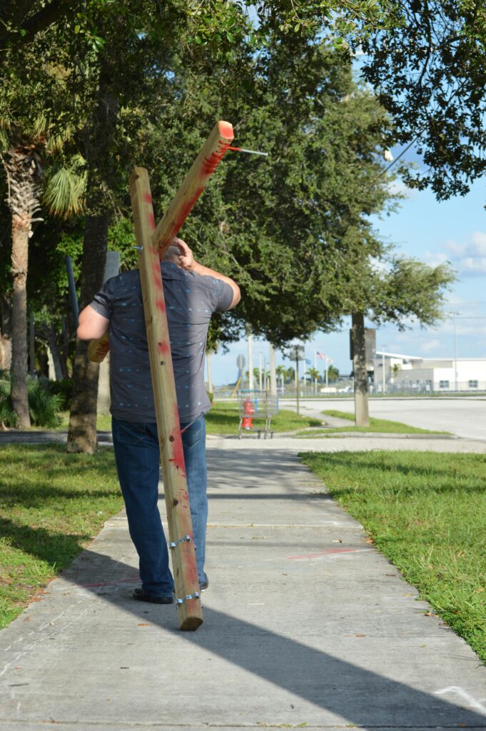A man walks on a sidewalk carrying a wooden cross, surrounded by green trees.
