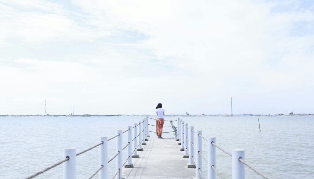 A woman stands quietly on a serene pier overlooking the calm ocean under a cloudy sky.