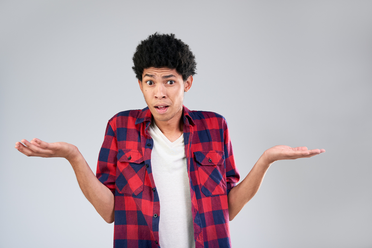 Young man shrugging with palms up, wearing a red plaid shirt over a white tee, looking surprised against a gray background.